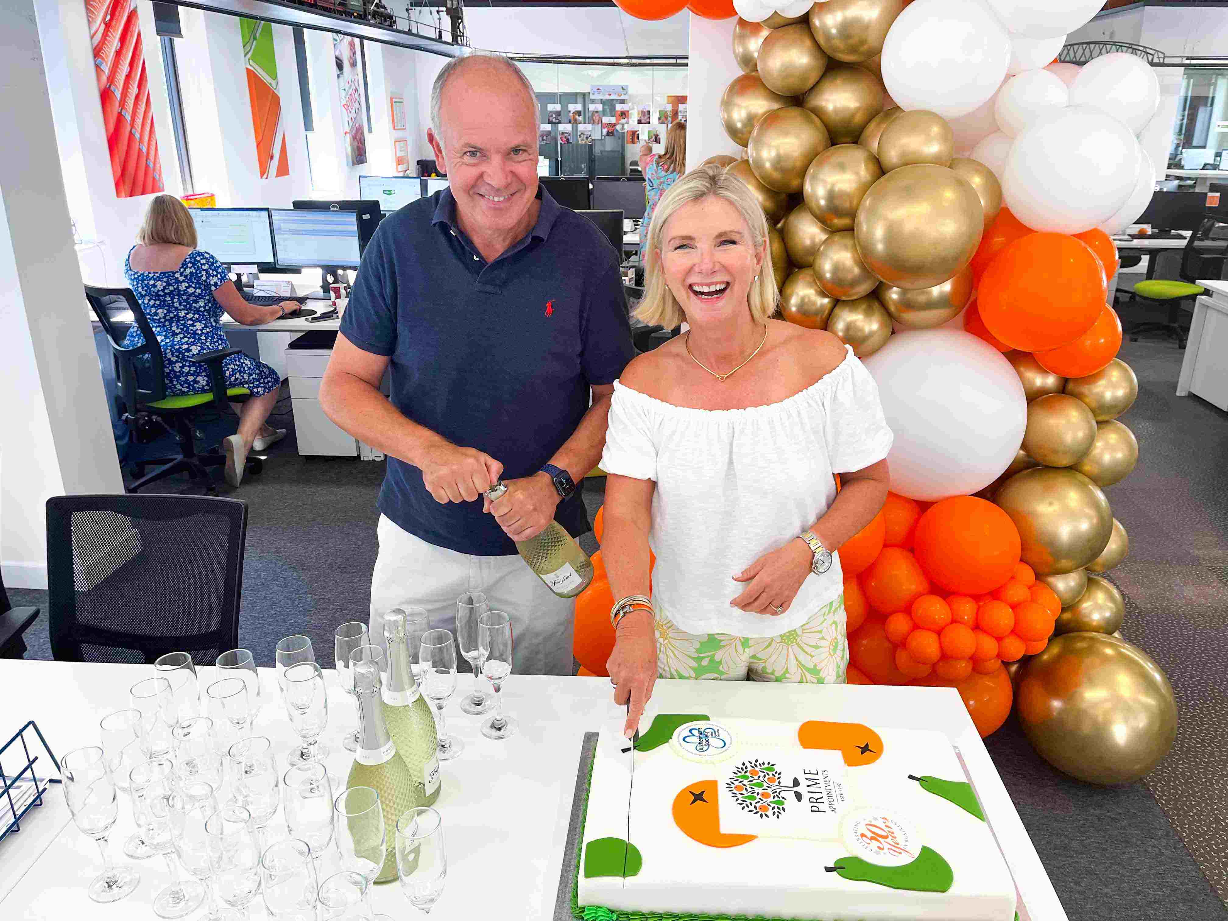 Robyn Holmes, MD & Peter Holmes, Director Cutting Cake in Prime Appointment 30th year celebrations with White, Gold & Orange Balloons in the background